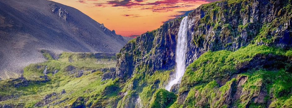 A stunning waterfall flowing down a cliffside with greenery and mountains in the background during sunset