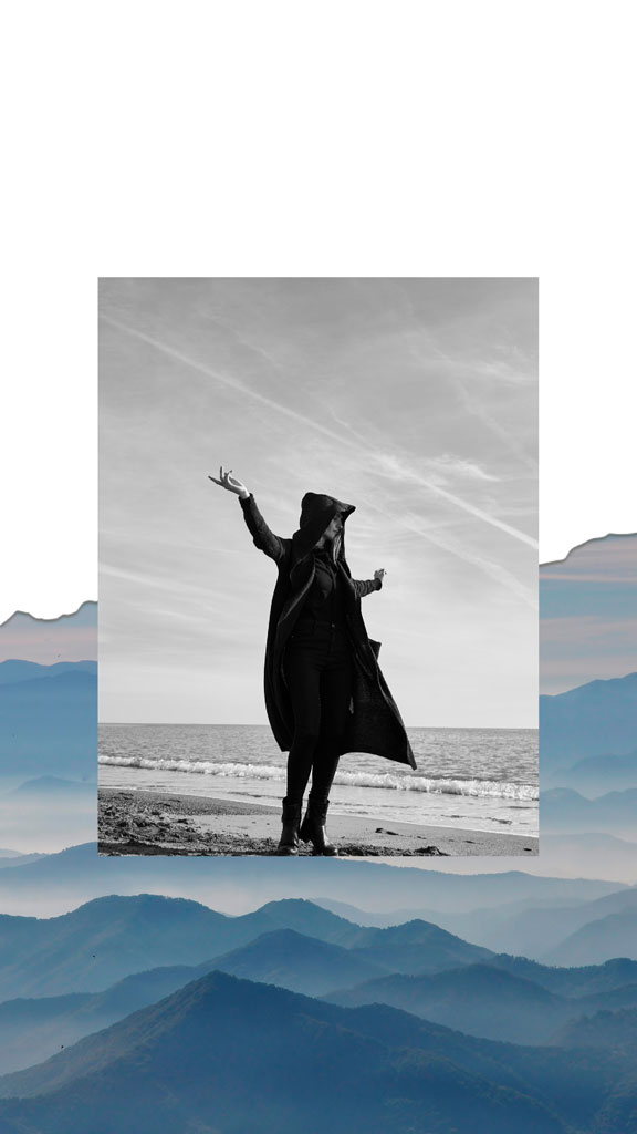 A black and white image of a woman in a long coat standing on a beach with her arms outstretched.