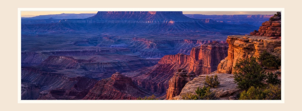 Wide desert canyon landscape with layered red cliffs and a pale sky framed by a beige border.