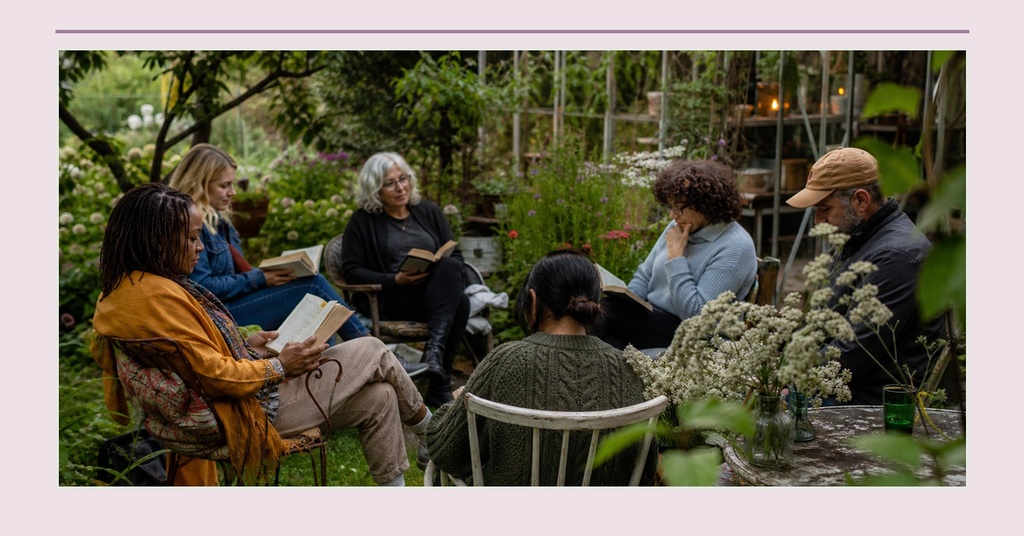 People seated outdoors in a garden, holding books and chatting among flowers and greenery.