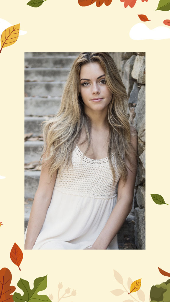 A young woman with long blonde hair wearing a white dress leaning against a stone wall surrounded by autumn leaves.