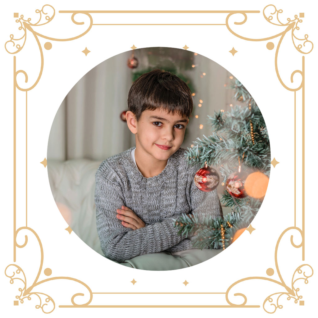 A young boy in a grey sweater sitting by a Christmas tree with ornaments
