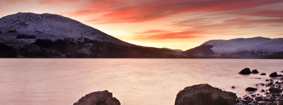 Snow-capped mountains and a calm lake at sunset with orange and pink hues in the sky.