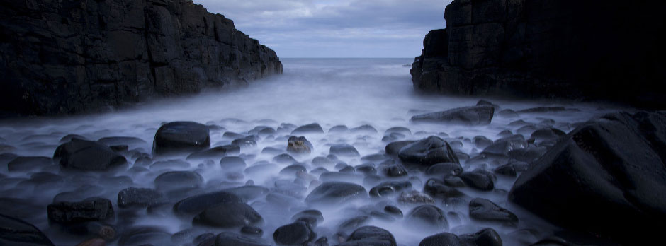 A long exposure photograph of a rocky beach at dusk with water flowing around the rocks.